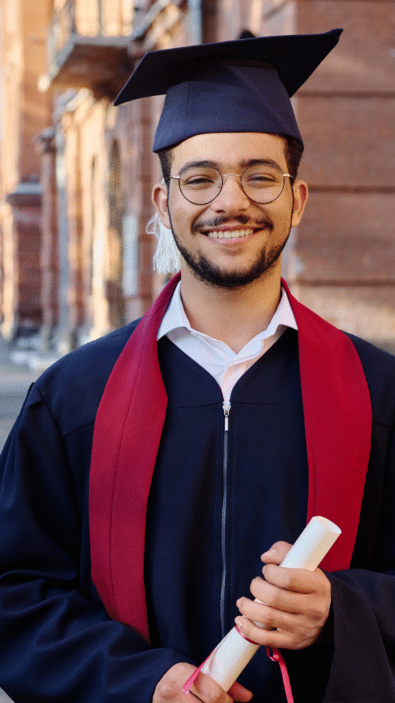 A graduate in a black gown and cap holds a rolled diploma tied with a red ribbon, posing with a proud expression against a yellow background.